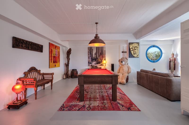 A spacious living room with a pool table, a large sofa, a wooden bench, and various decorative items including paintings, a statue, and a teddy bear. The room features a hanging pendant light and a colorful rug.