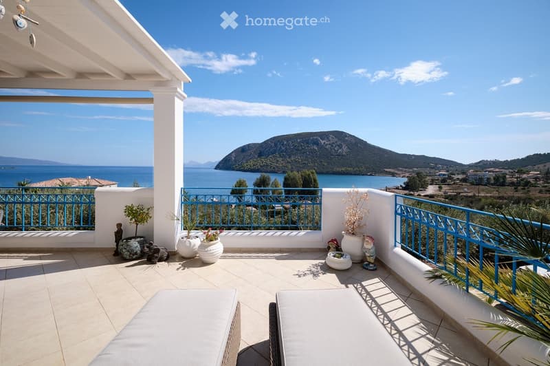 A spacious balcony with a tiled floor, white walls, and a blue metal railing, overlooking a scenic sea view with mountains in the distance. Decorated with potted plants, a small table, and a garden gnome.