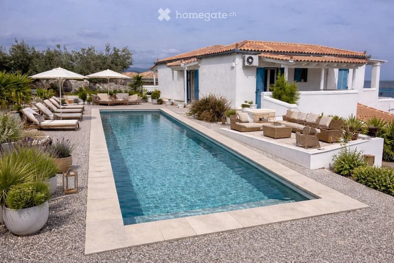 A modern outdoor area featuring a rectangular swimming pool surrounded by lounge chairs, umbrellas, and potted plants. The area is bordered by gravel and includes a white house with blue shutters in the background.