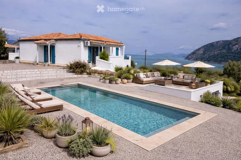 Outdoor area with a swimming pool, lounge chairs, sofas, coffee tables, and umbrellas, overlooking the sea with surrounding plants and a white house with blue shutters.