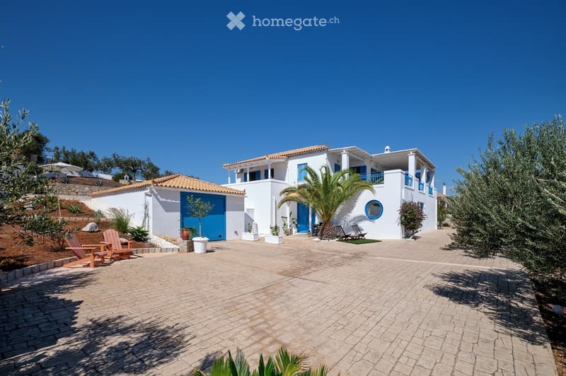 Two-story white house with blue accents, terracotta roof, and a spacious brick-paved driveway. Features include a garage, outdoor seating area, and surrounding greenery.