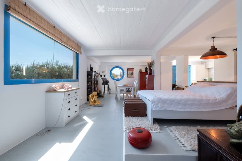Bright, modern bedroom with white walls and ceiling, large window with blue frame, bed with white bedding, dresser, small table with chairs, decorative items including a vase, picture frame, and a red bowl.