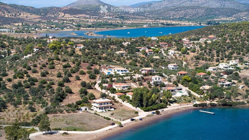 Aerial view of a coastal village with scattered houses, trees, and a lake in the background, surrounded by hills and mountains.