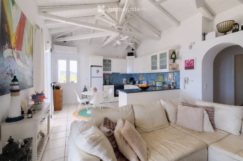 Bright, airy living and dining area with white walls, exposed wooden beams, and a high ceiling. Features a cream-colored sectional sofa, a small dining table with white chairs, a modern kitchen with white cabinets and stainless steel appliances, and vario