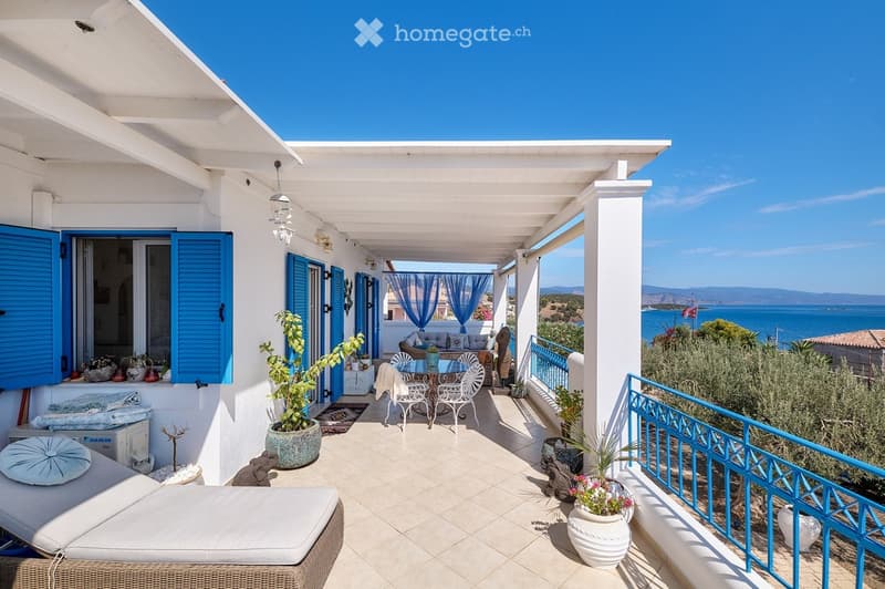 A spacious, covered balcony with a view of the sea, featuring a sofa, a round table with chairs, potted plants, and decorative cushions.