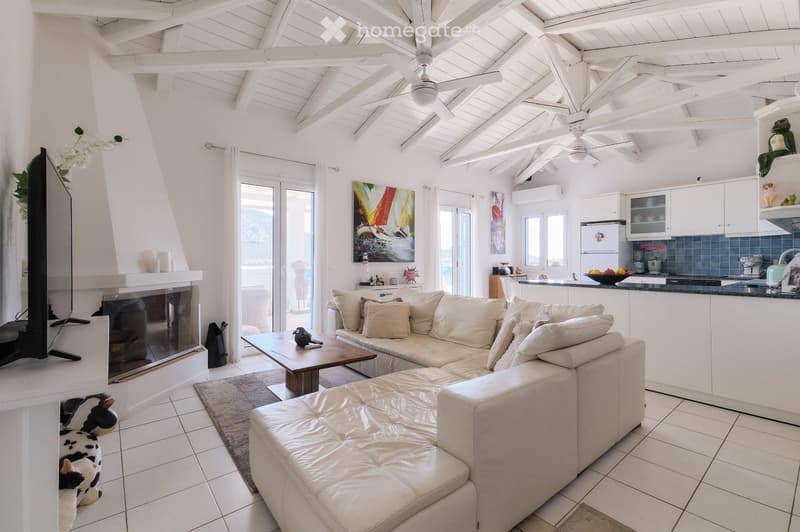 Open-plan living and dining area with a white leather sofa, coffee table, ottoman, fireplace, TV, and a modern kitchen with white cabinets and stainless steel appliances.