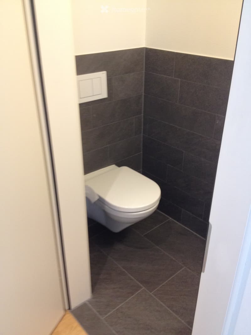 Modern bathroom with dark tiled walls and floor, white toilet, and a light switch on the wall.