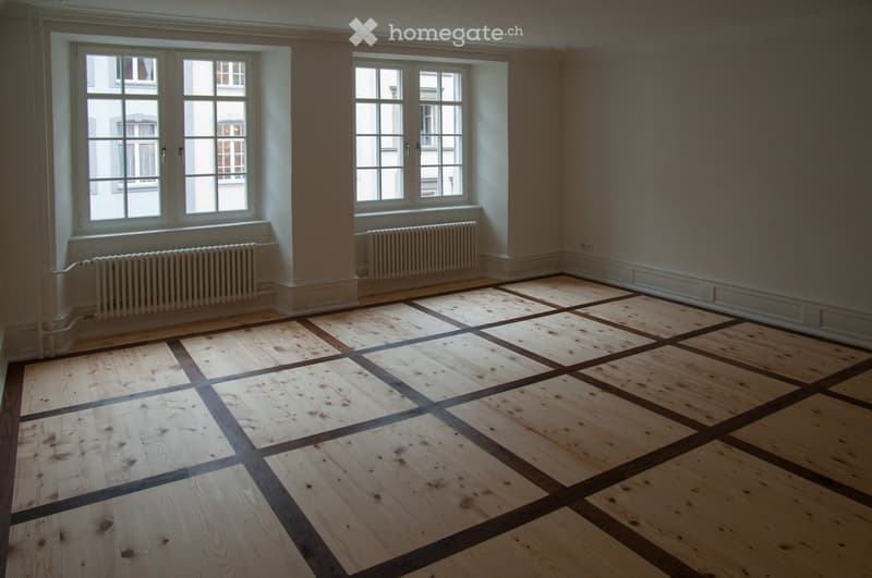Empty room with wooden floor and large windows, featuring radiators under the windows.