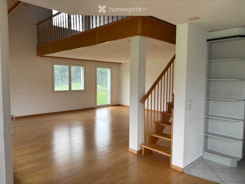 Bright, open-plan living space with wooden flooring, large windows, and a staircase leading to an upper level. The room is completely empty, with no furniture or decorations.