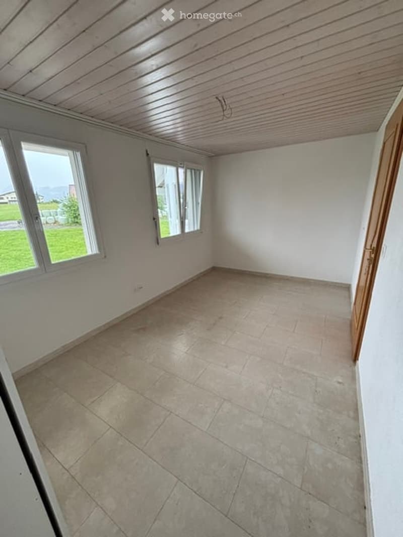 Empty room with tiled floor, white walls, and large windows offering a view of a green outdoor area.