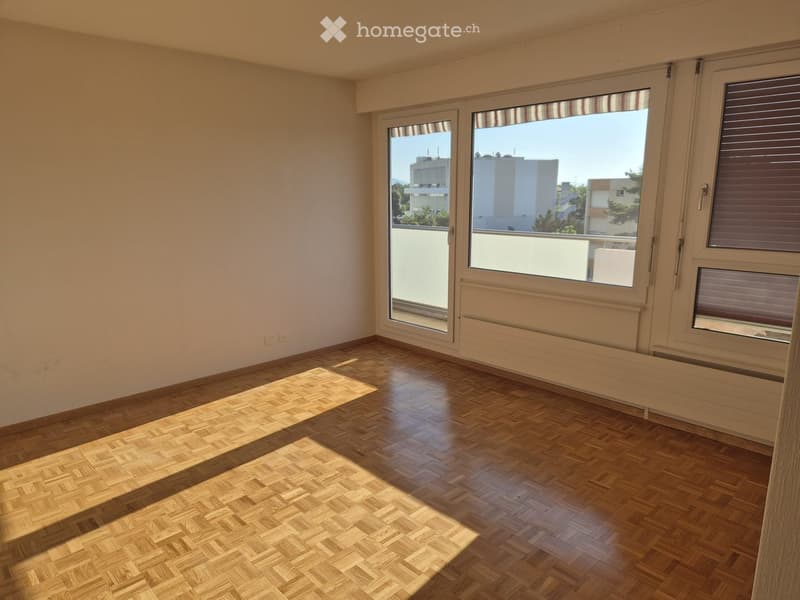 Empty room with wooden parquet flooring, large windows with blinds, and a sliding glass door leading to a balcony.