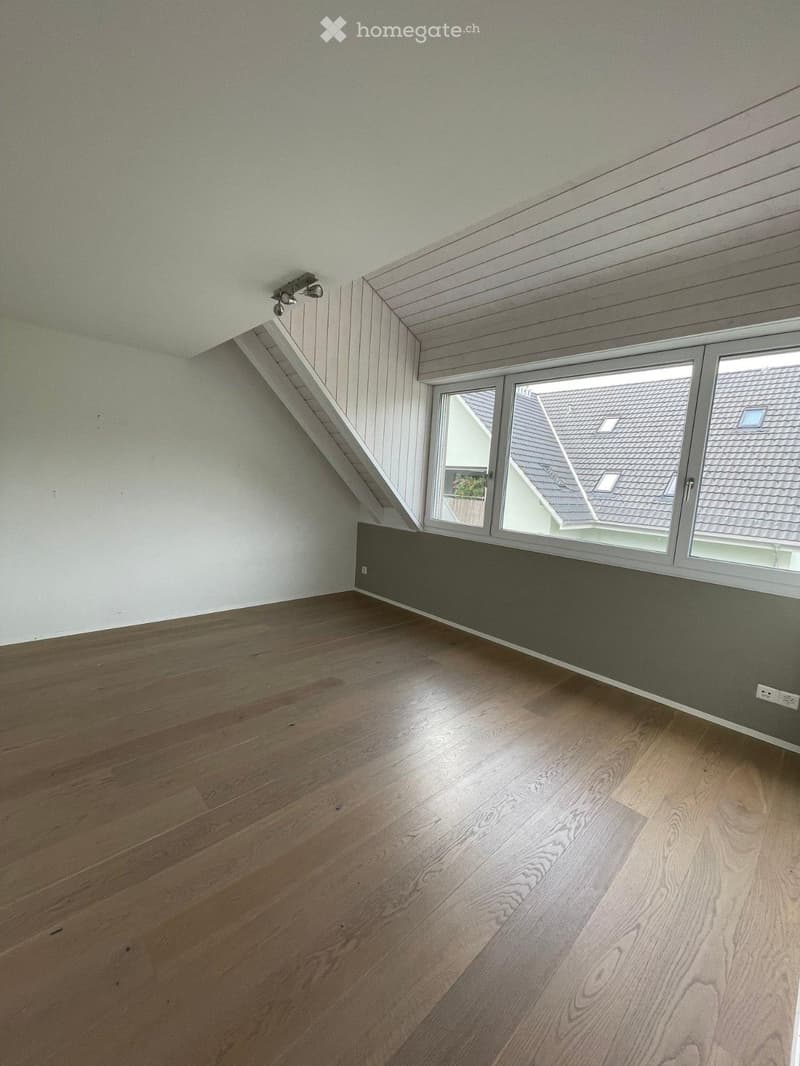 Empty attic room with wooden flooring, white walls, and large windows offering a view of neighboring rooftops.