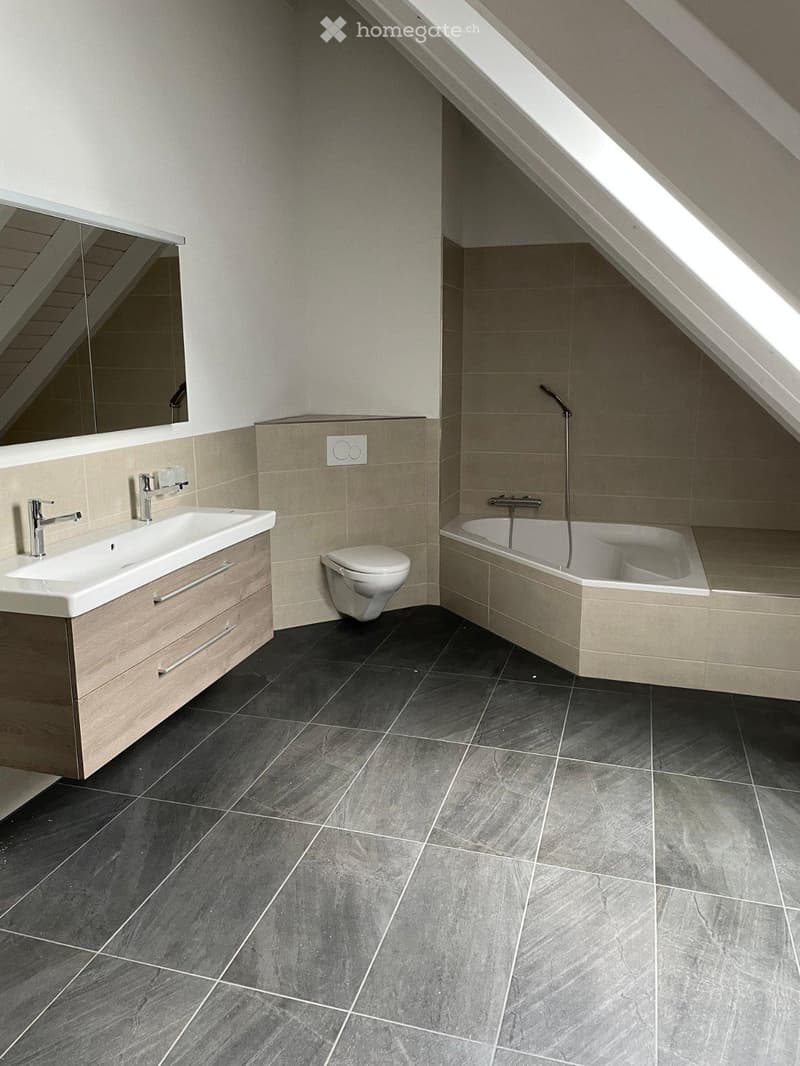 Modern bathroom with dark grey tiles, a white sink with wooden cabinet, a toilet, and a bathtub with a handheld showerhead.