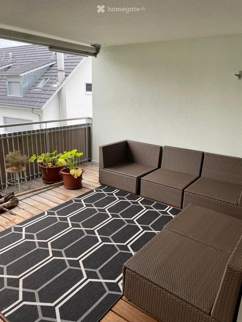 Balcony with wooden decking, black and white geometric rug, wicker sofa and chairs, small table with potted plants, and a view of neighboring building.