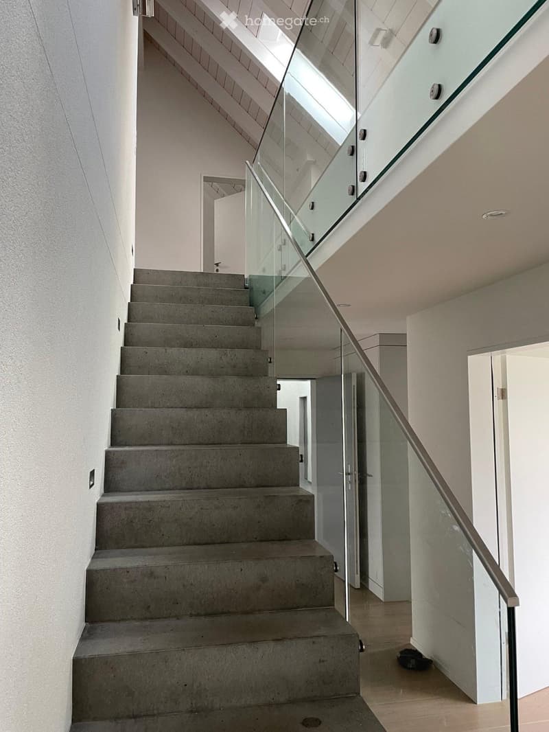 Modern staircase with concrete steps and a sleek glass railing, leading to an open area with white walls and a wooden floor.
