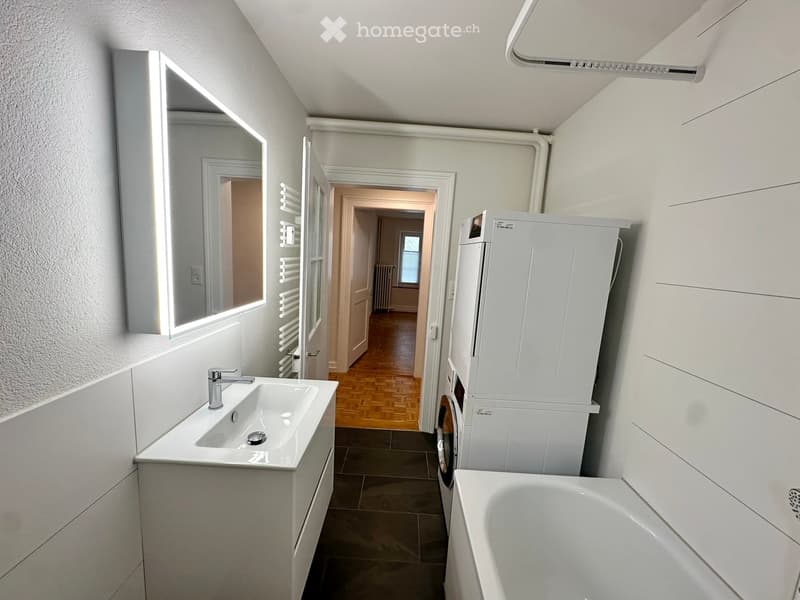 Modern bathroom with a white sink, bathtub, mirror, and washing machine. The space features a wooden floor and white walls.