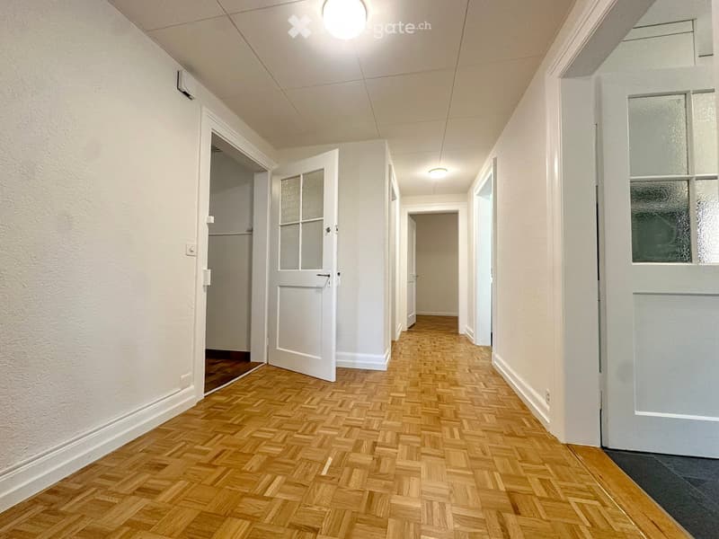 A long, narrow hallway with wooden herringbone flooring, white walls, and several doors with frosted glass panels. The ceiling has recessed lighting and visible air conditioning vents.