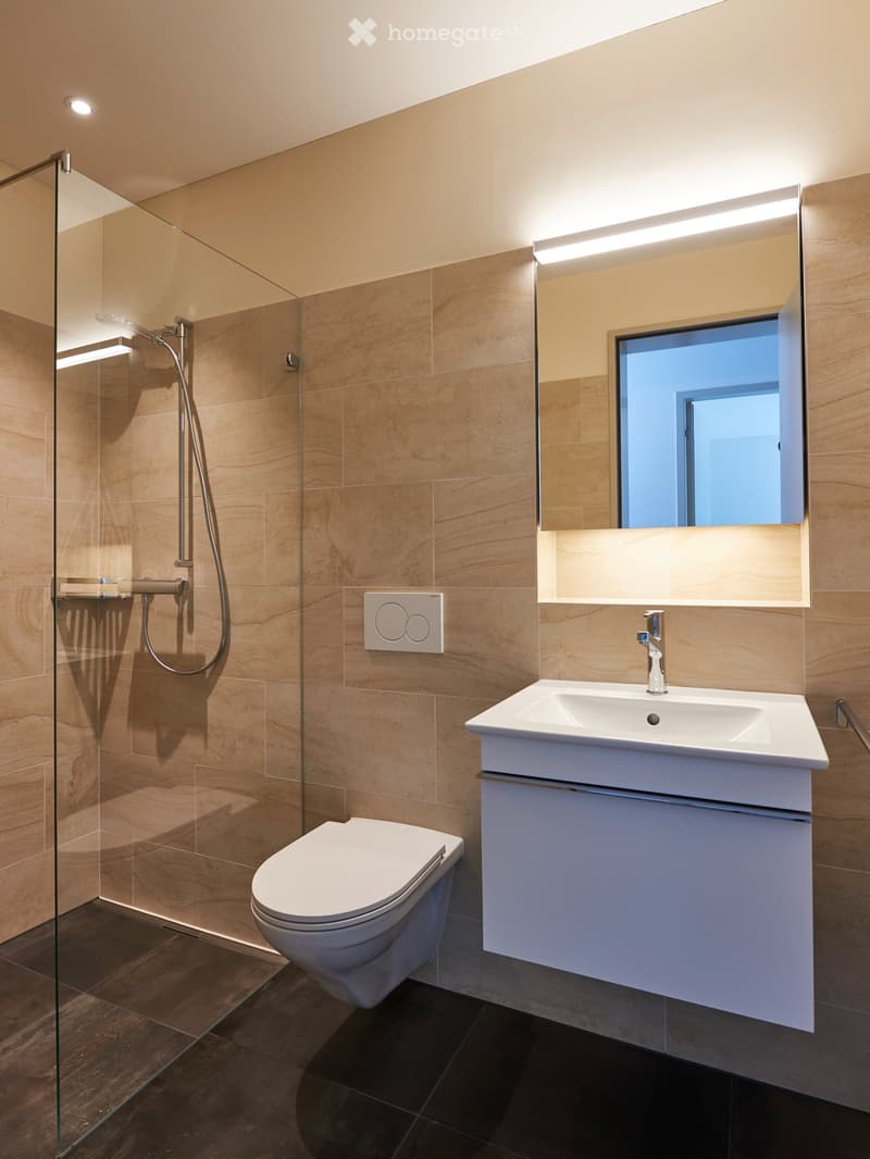 Modern bathroom with beige tiles, a white toilet, a white sink with a chrome faucet, a large mirror with built-in lighting, and a glass-enclosed shower with a handheld showerhead.
