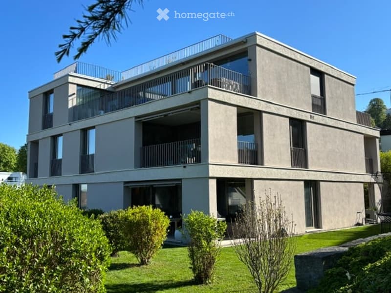 Modern three-story building with large windows and balconies, surrounded by a well-maintained lawn and shrubs.