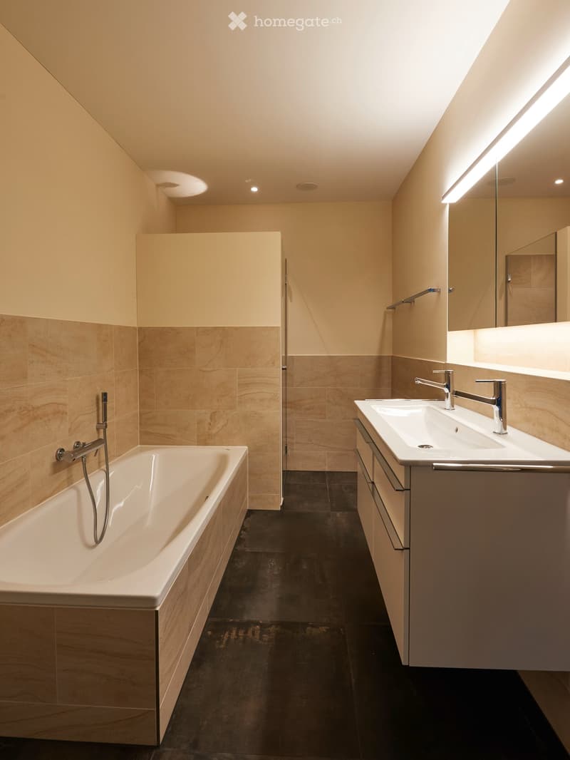 Modern bathroom with a bathtub, double sink, and large mirror illuminated by LED lighting. Beige tiles and dark flooring.