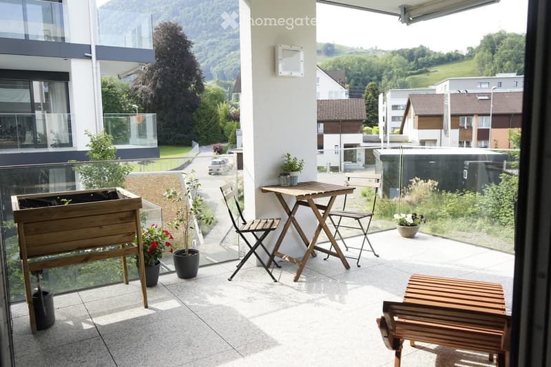 Balcony with a small wooden table, two chairs, a bench, and potted plants, overlooking a scenic landscape with hills and houses.