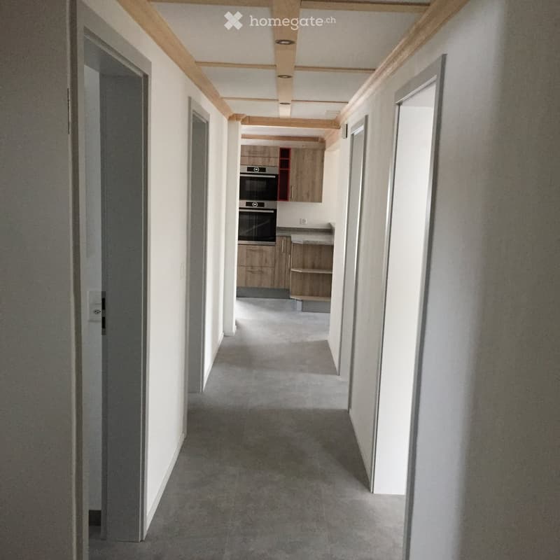 Empty hallway with white walls, grey carpet, and wooden beams on the ceiling. A kitchen area is visible at the end with cabinets and appliances.