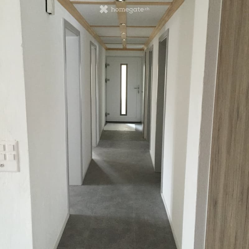 Clean, modern hallway with white walls, grey carpeted floor, and wooden ceiling beams. Doors line both sides, with a small rug placed near the far end.