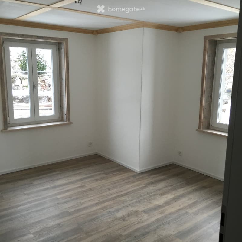 Empty room with light-colored wooden flooring, white walls, and two windows with wooden frames. The ceiling features exposed wooden beams.