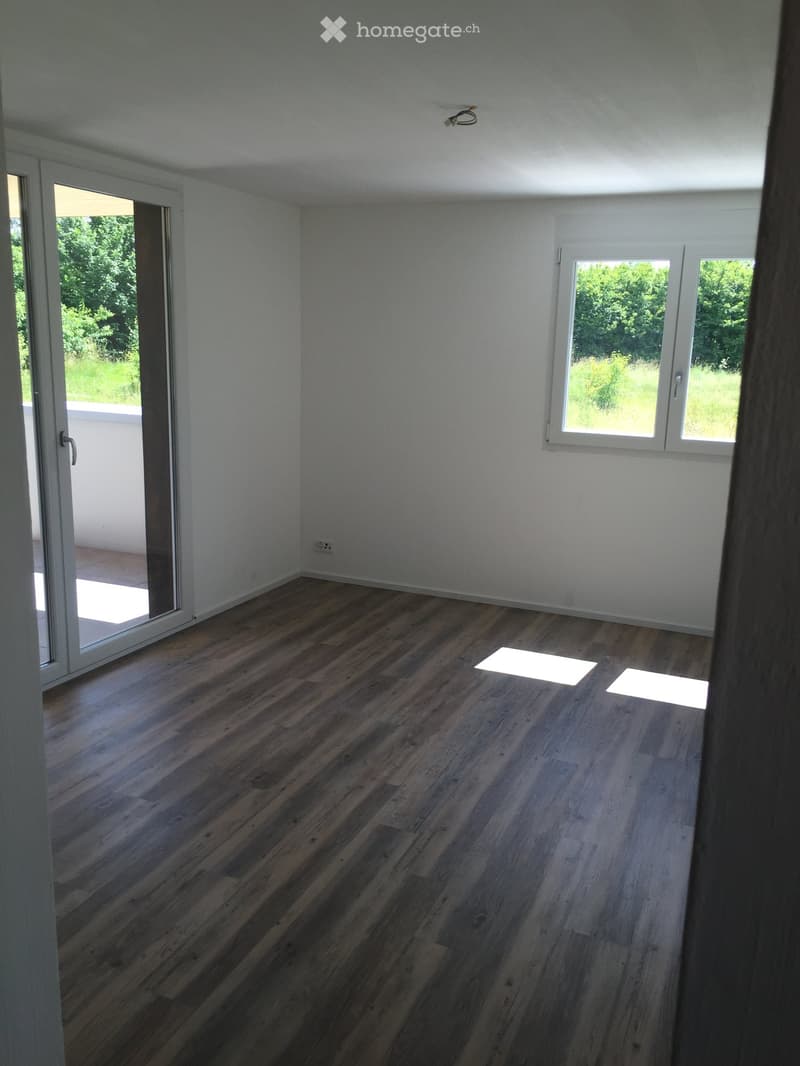 Empty room with wooden flooring, white walls, large windows, and a sliding glass door leading to a balcony.