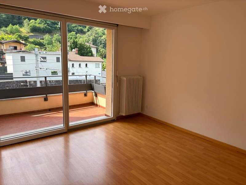 Empty room with wooden flooring, large sliding glass doors leading to a balcony with a view of greenery and buildings.