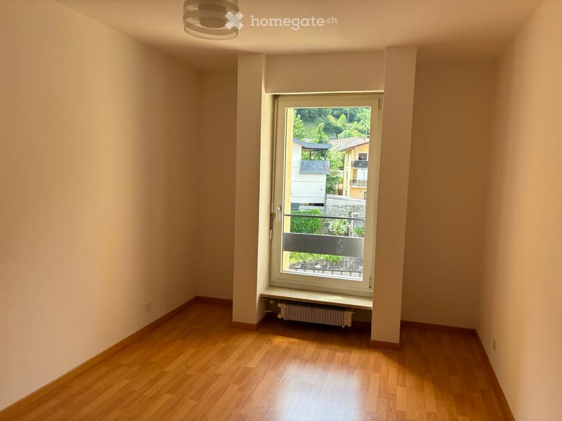 Empty room with wooden flooring, white walls, and a large window with a view of greenery and neighboring buildings.