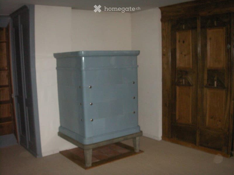 {"visible_features":[],"type":"Room","description":"The image shows a small, square, gray-colored cabinet or storage unit placed on a wooden platform or stand in a room with white walls and a wooden wardrobe or cabinet visible in the background."}