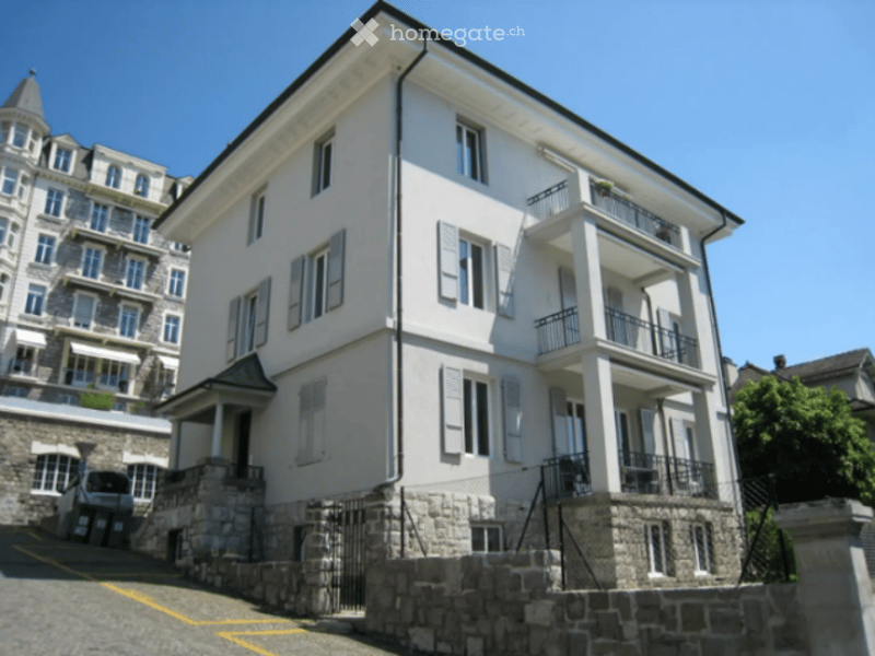 A multi-story residential building with white walls, multiple balconies, and stone base. The building features classic European architecture with shutters and a gated entrance.