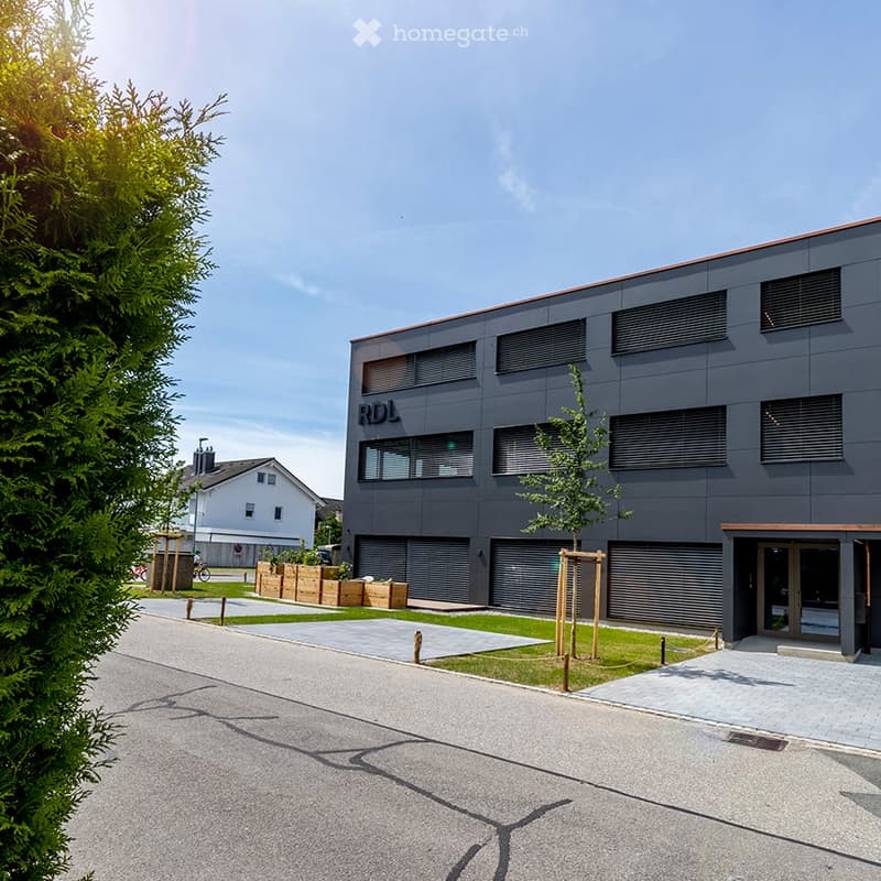 Modern two-story building with dark gray facade, large windows, and a small landscaped area in front.