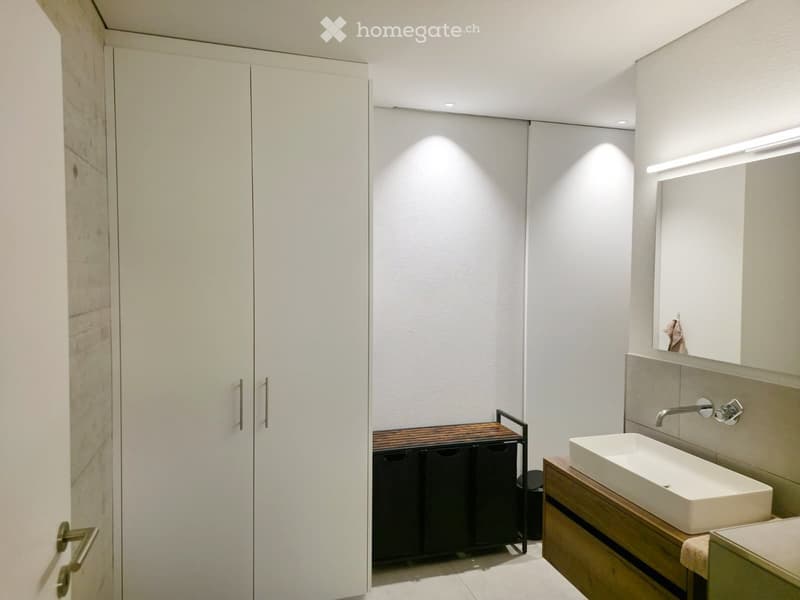 Modern bathroom with white walls, a sink on a wooden vanity, a large mirror, and a white cabinet with silver handles.