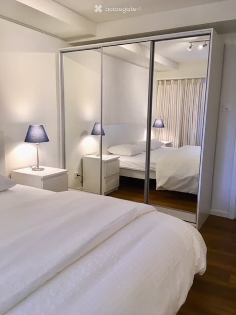 Modern bedroom with a large bed, nightstand, lamp, and a mirrored wardrobe. The room features wooden flooring and soft lighting.