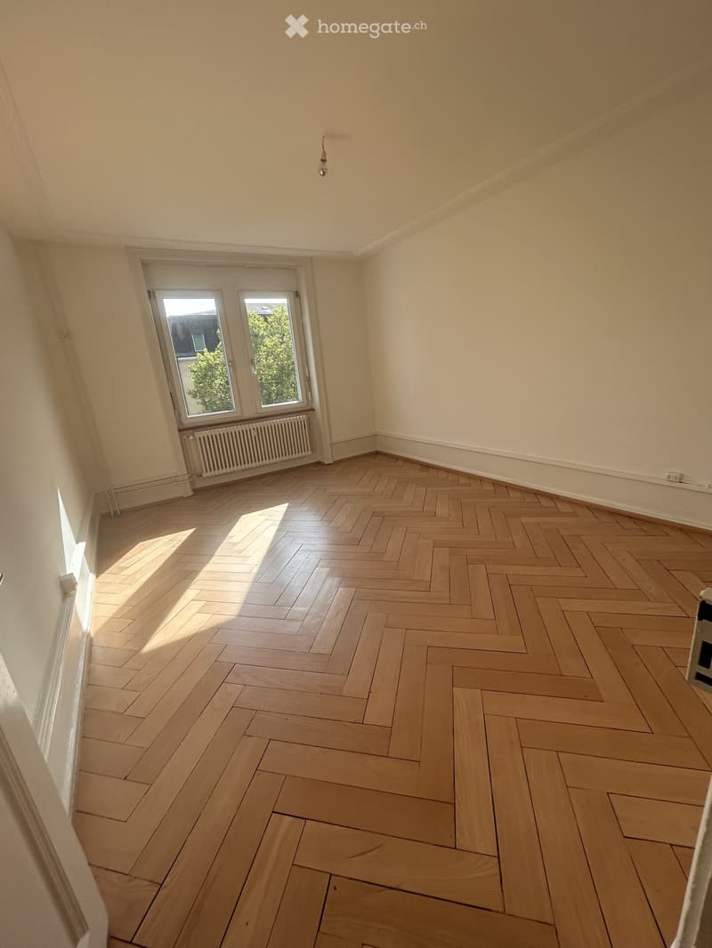 Empty room with light wooden herringbone flooring, white walls, and a window with a radiator below it.
