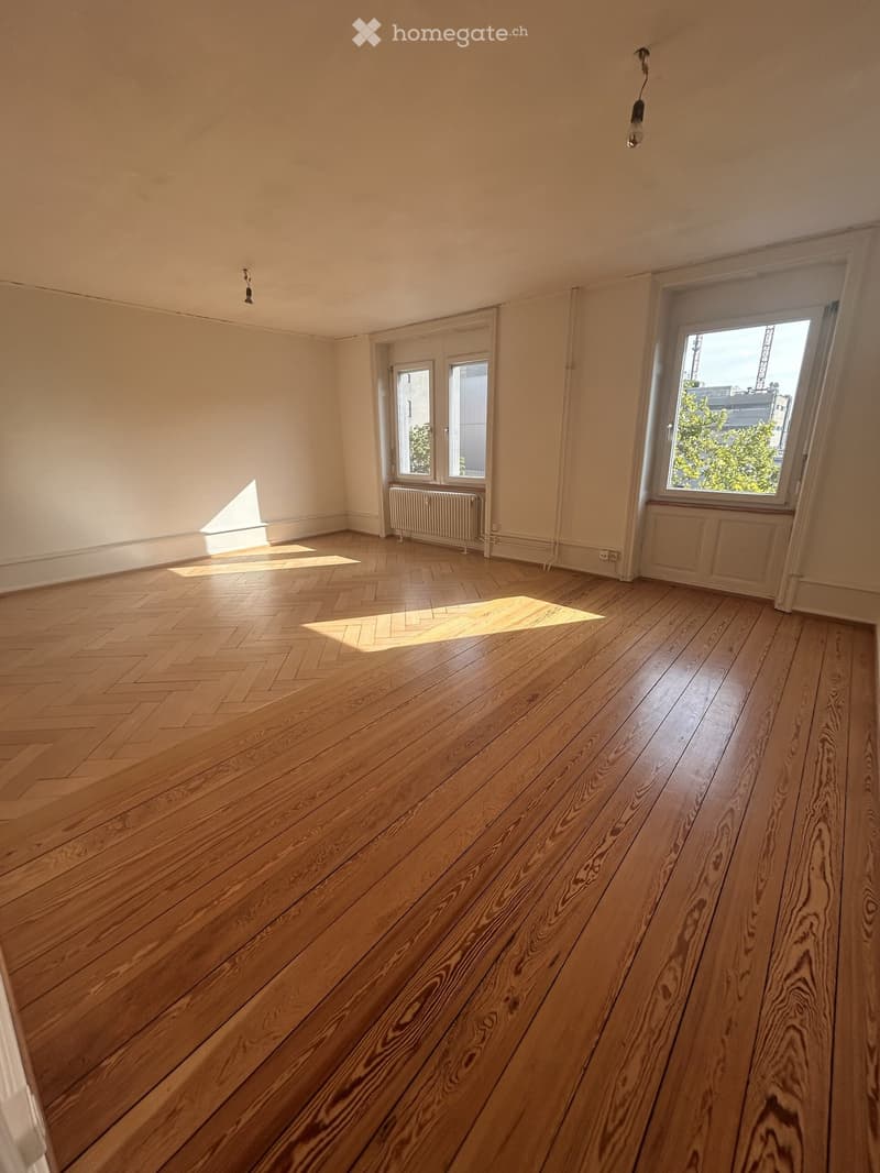 Empty room with wooden herringbone flooring, white walls, and large windows allowing natural light. Radiators are visible under the windows.