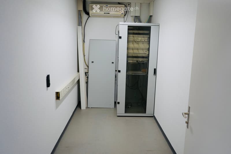 Empty room with white walls and a tiled floor, containing a server rack, an air conditioning unit, and electrical panels.