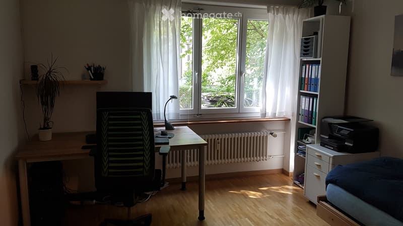 Well-lit room with wooden flooring, large window with white curtains, desk with chair, screen, lamp, shelf with binders, printer, and a small bed.