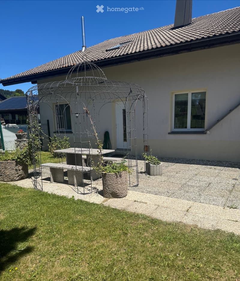 Garden area with a stone table, benches, a decorative metal arch, and potted plants near a house.