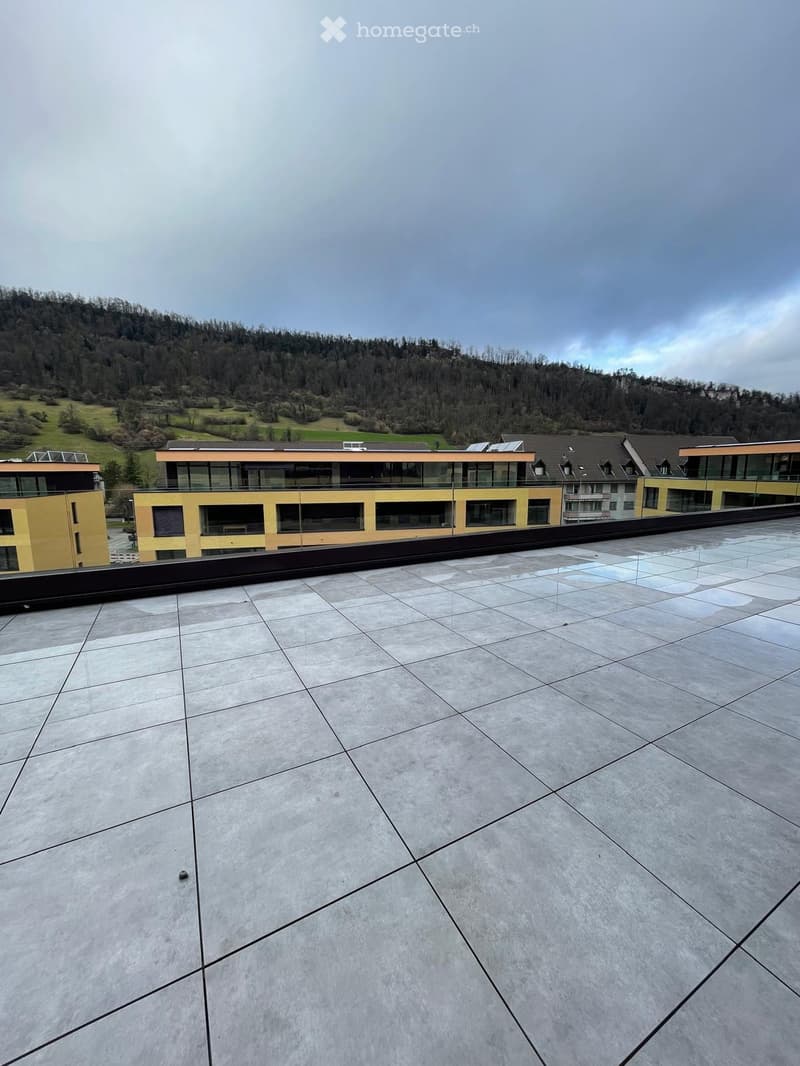 Roof terrace with large, light-colored tiles and a modern, sleek design. The terrace overlooks a modern building complex with yellow and orange facades and large glass windows, set against a backdrop of green hills and a partly cloudy sky.