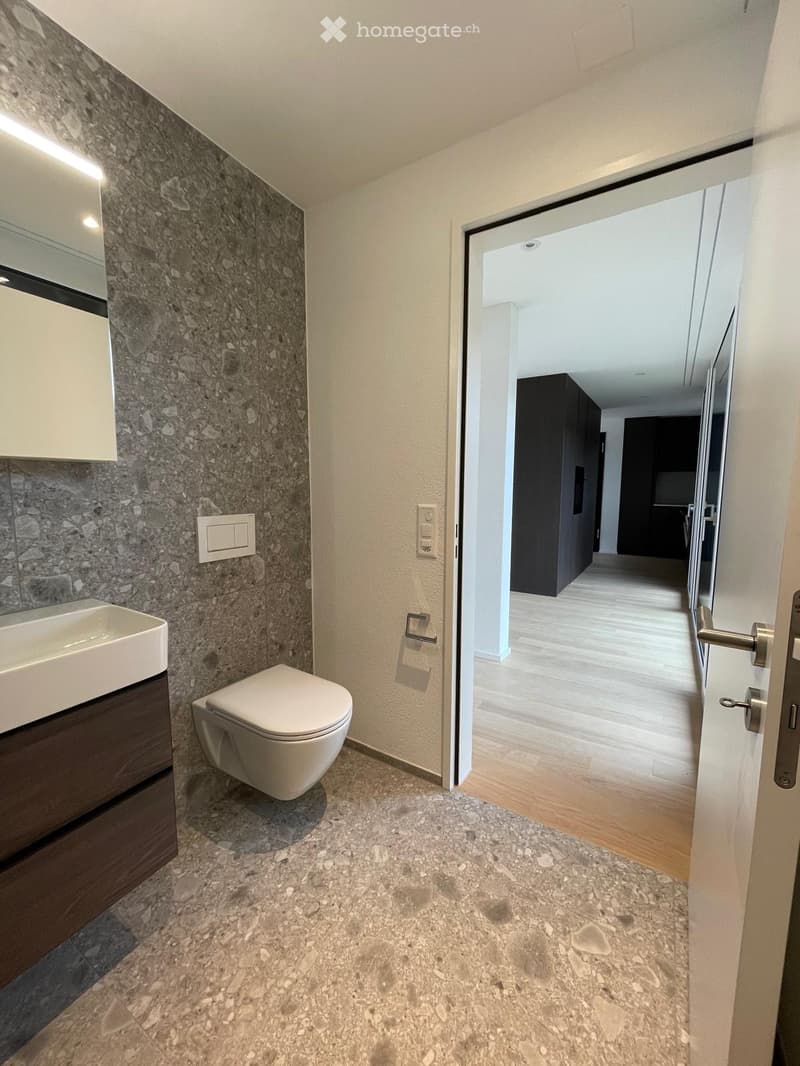 Modern bathroom with a white sink, toilet, and dark wooden cabinet. The walls feature a textured stone design, and the floor is made of polished stone tiles. An open doorway leads to a hallway with wooden flooring.