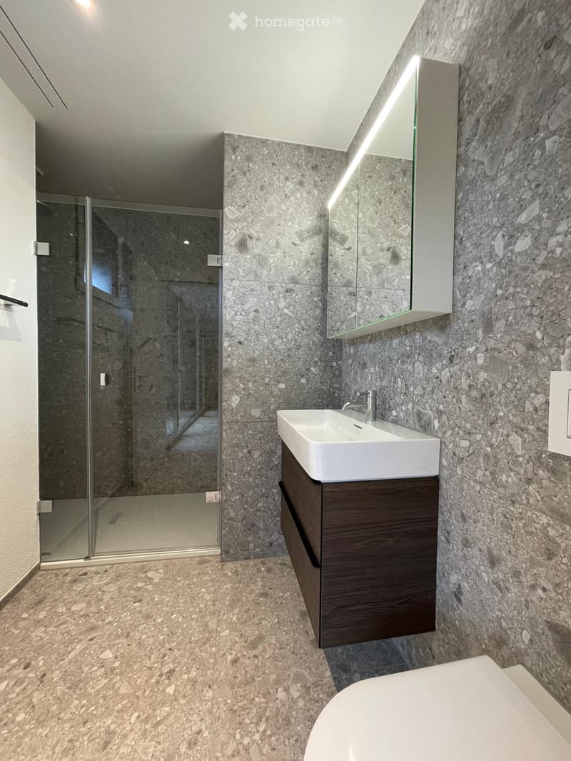 Modern bathroom with marble walls and floor, white sink with dark wooden cabinet, large mirror, and glass-enclosed shower area.
