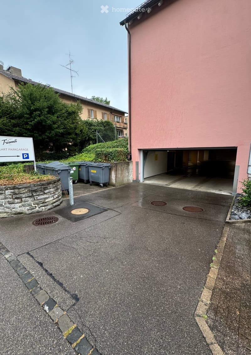 A paved driveway leading to a garage entrance of a pink building, with a sign indicating parking directions and several trash bins nearby.