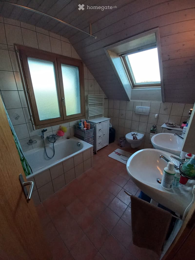 Bathroom with a bathtub, two sinks, toilet, cabinet, and a window with blinds. The room features tiled walls and a slanted ceiling typical of an attic space.