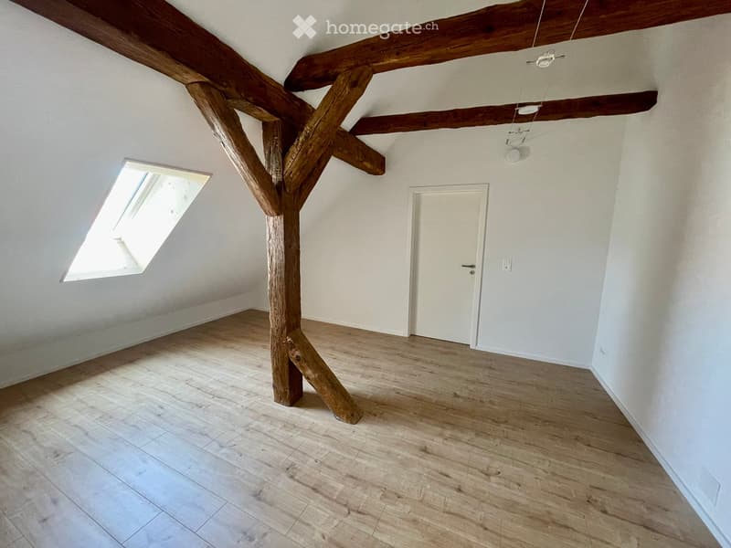Empty room with wooden floor, exposed wooden beams, skylight window, and white walls.