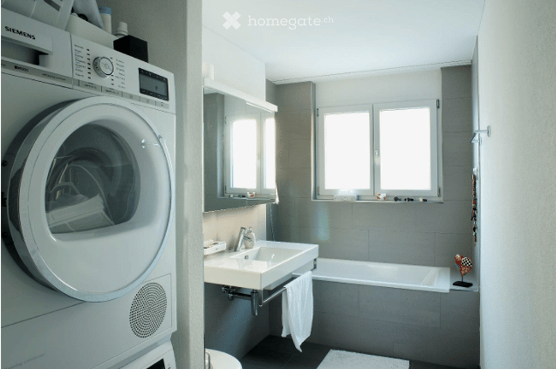 Modern bathroom with a white sink, chrome faucet, bathtub, mirror, and a white washing machine. The space features grey tiled walls and a small window above the sink.