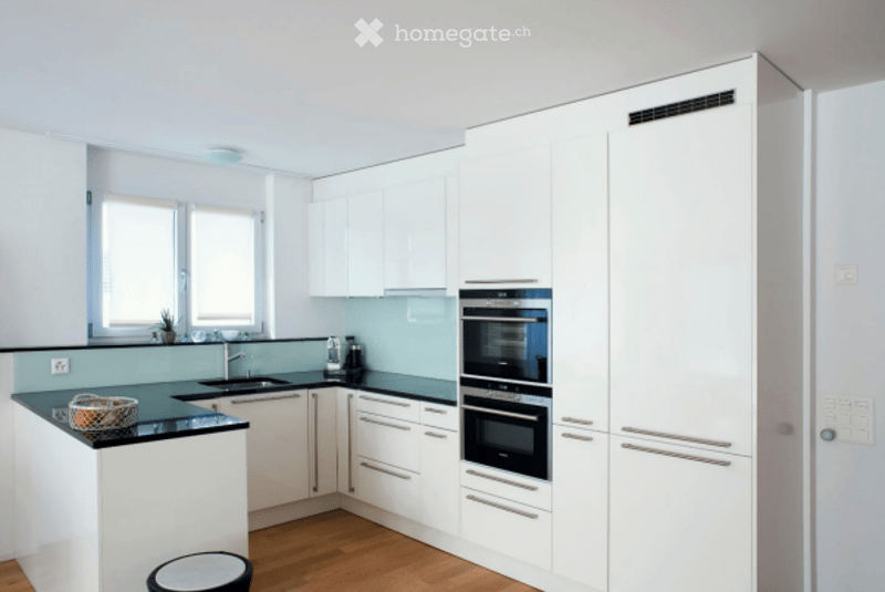 Modern kitchen with white cabinets, black countertops, and stainless steel appliances including an oven and fridge. Features a sink with a faucet, a small stool, and a window with a plant on the windowsill.