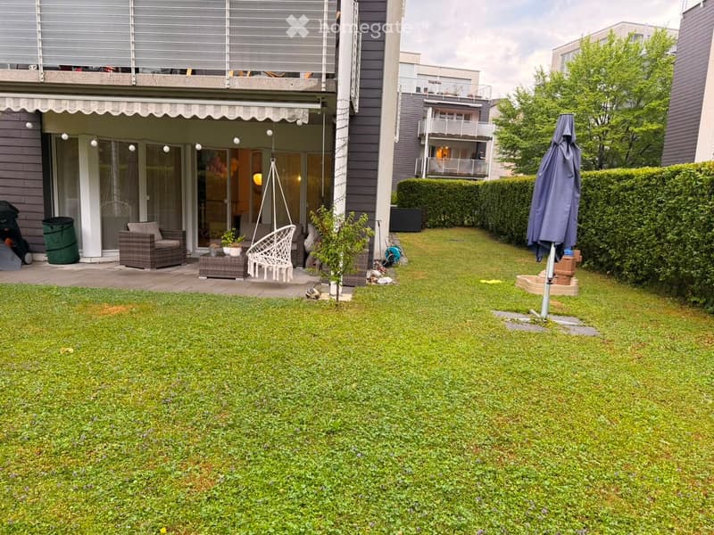 Garden with green lawn, patio area with wicker furniture, swing chair, umbrella, and trash bin.